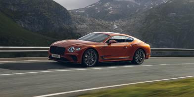 Side angled view of Bentley Continental GT Speed in Orange Flame, driving along highway with mountains in backdrop. 