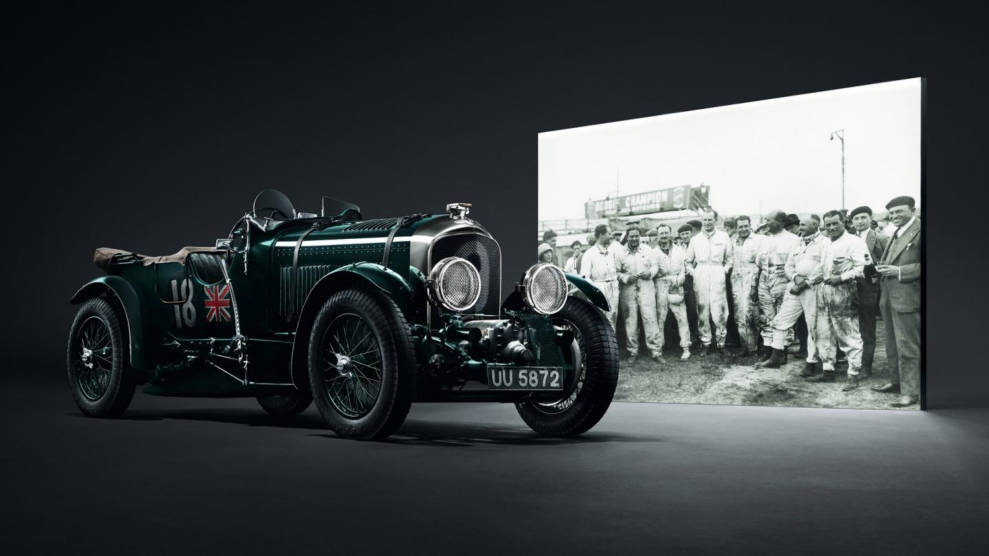 Front three quarter of the Tim Birkins Bentley Blower in front of a group shot of all of the Bentley Boys