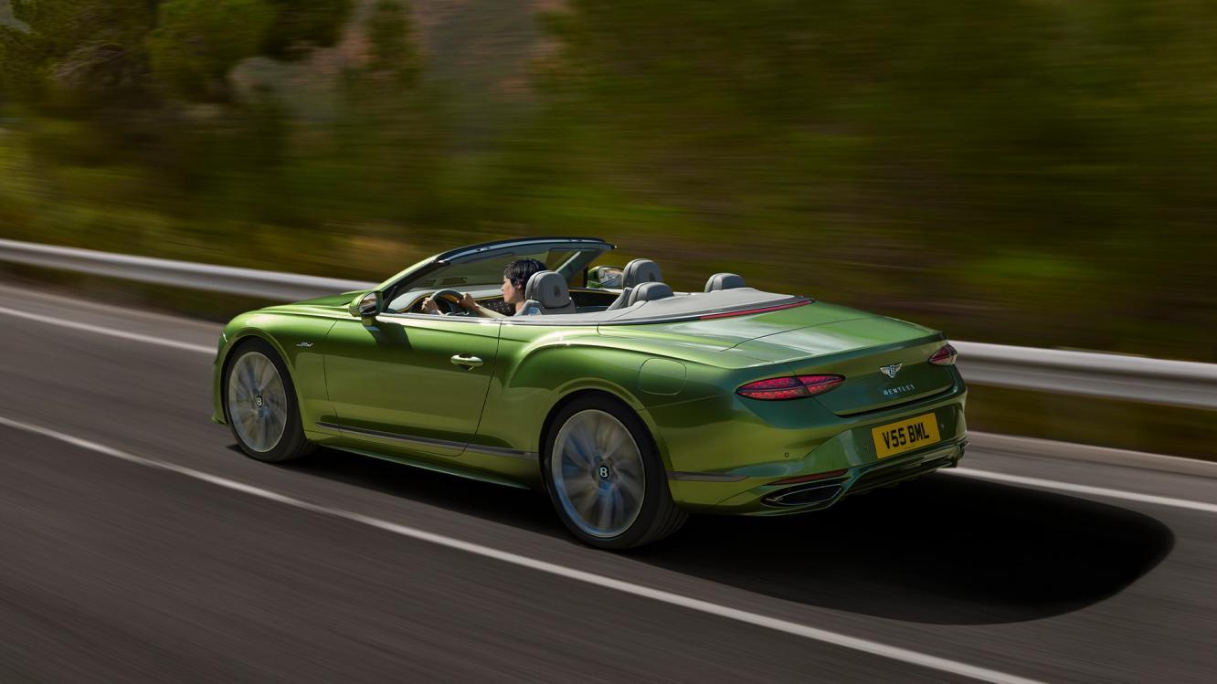 Rear side angled view of the new Bentley Continental GT Convertible Speed in Tourmaline Green featuring 21 inch directional and diamond cut Speed wheels driving in a rural environment
